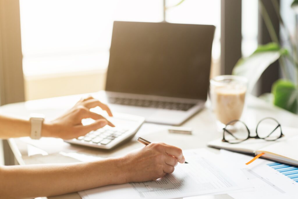A person uses a calculator and pen to work on documents at a desk with a laptop, glasses, and a drink.