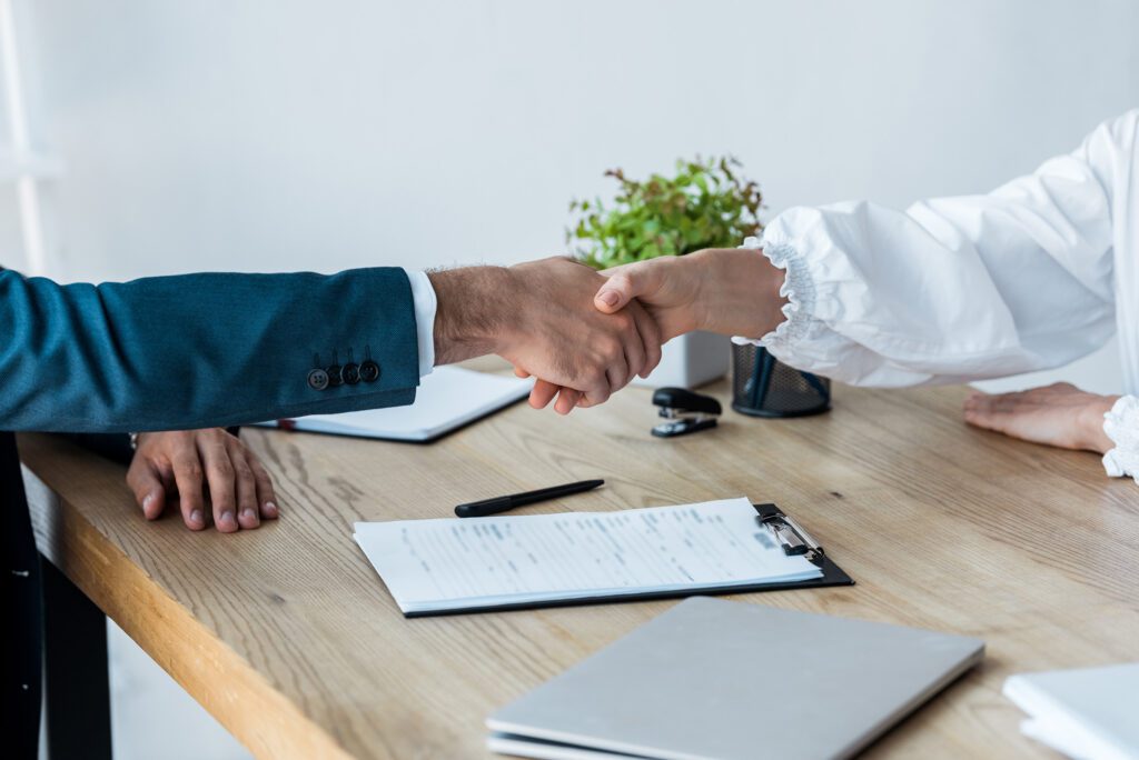 Two people shake hands across a wooden desk with paperwork, a clipboard, pen, and potted plant visible in the background.