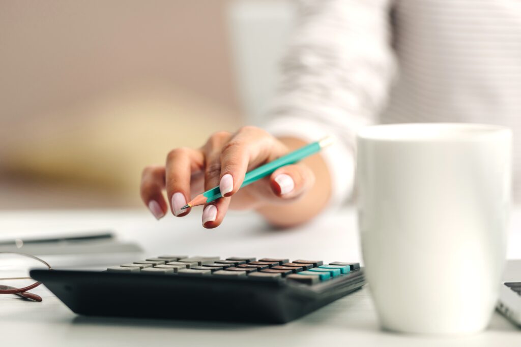 A person uses a calculator with one hand while holding a pencil, with a white coffee mug in the foreground.