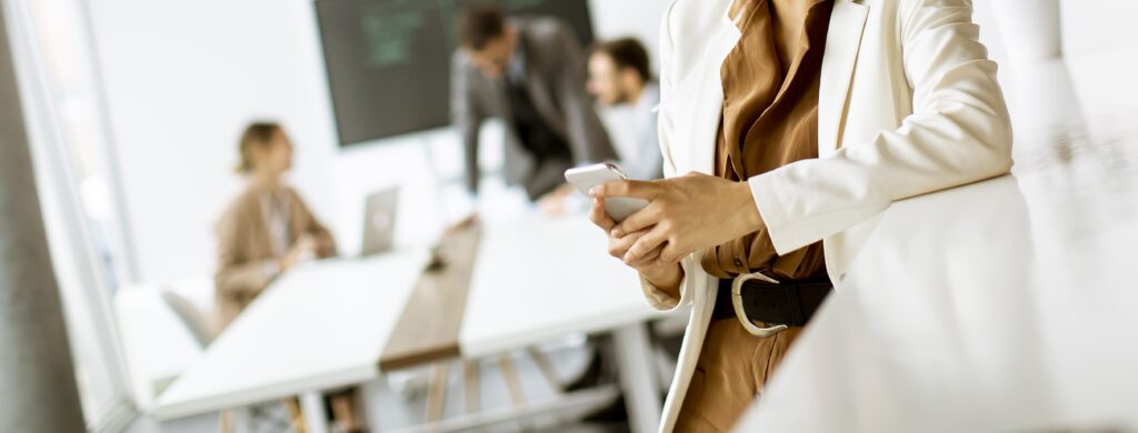 A person in business attire holding a smartphone stands in the foreground, while three colleagues work and converse around a conference table in the background.