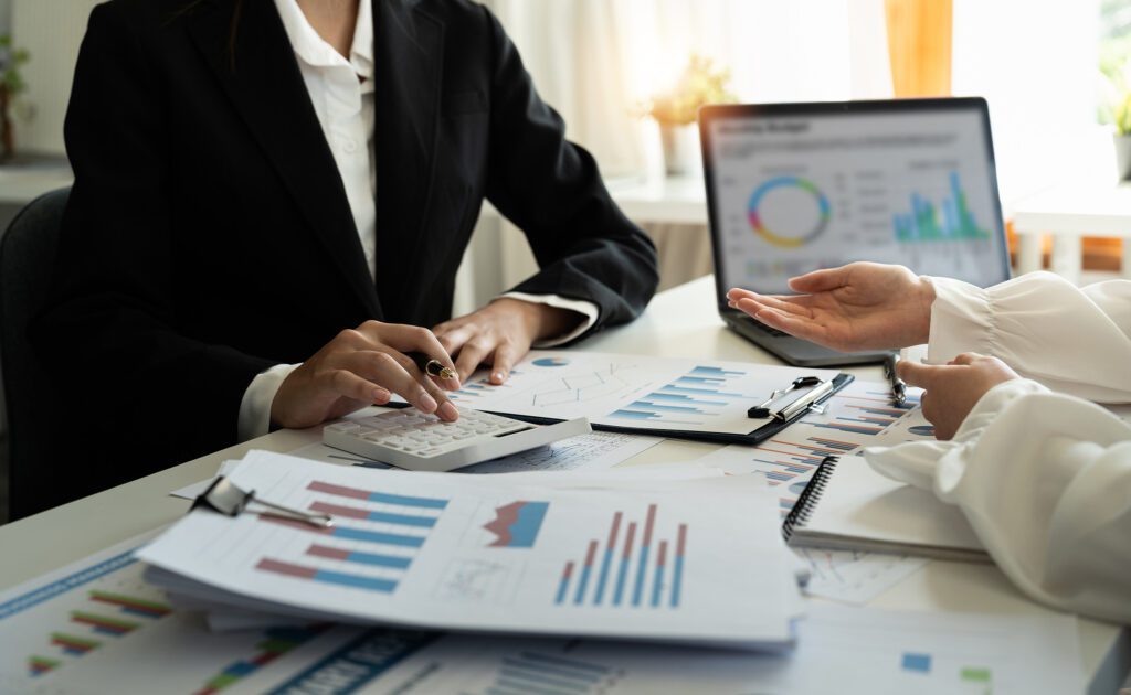 Two people in formal attire discuss financial charts and graphs at a desk with a laptop, documents, and a calculator.