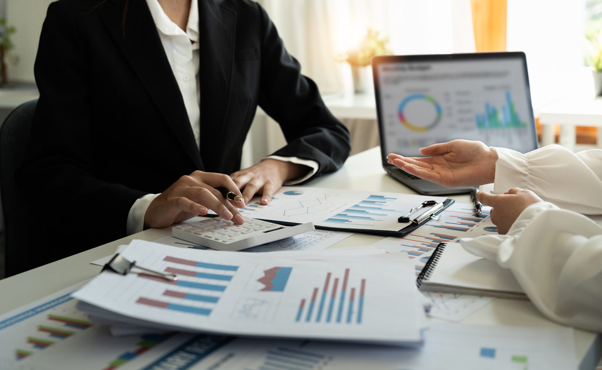 Two people in formal attire discuss financial charts and graphs at a desk with a laptop, documents, and a calculator.