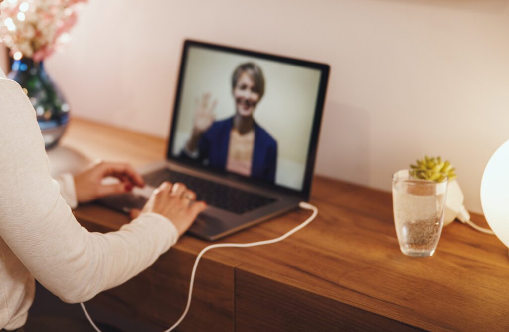 Person sitting at a desk using a laptop, with a video call on screen showing another person waving; a glass of water and lamp are also on the desk.